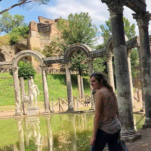 A student stands at a historical site, with a set of arches around a pool of water and a statue in the background