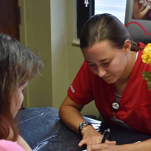 A nursing student writes on a piece of paper across a desk from a woman